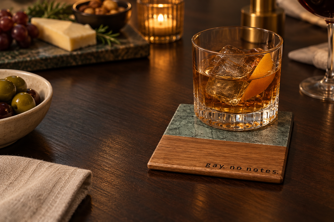 Whiskey glass on a green marble and wood coaster engraved “gay. no notes.” on a warmly lit table with olives and cheese.
