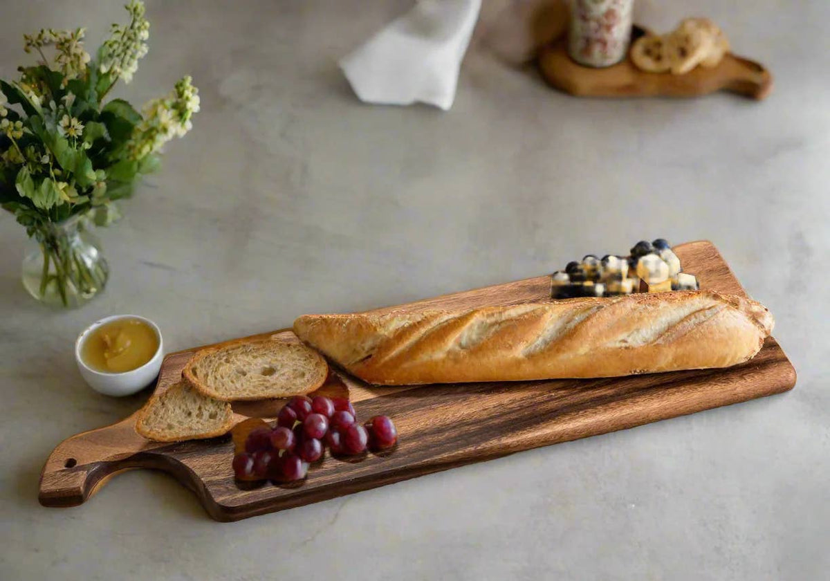 Extra long acacia serving board with handle topped with a baguette, grapes, and olives