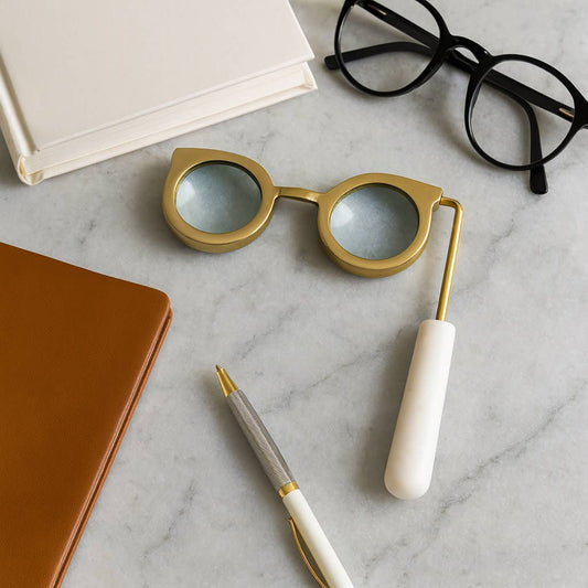 Gold eyeglass-shaped magnifying glass with white marble handle on a marble desk beside reading glasses and a notebook