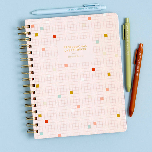 Blush grid spiral notebook with colorful confetti squares, Professional Overthinker, beside pens on a blue surface