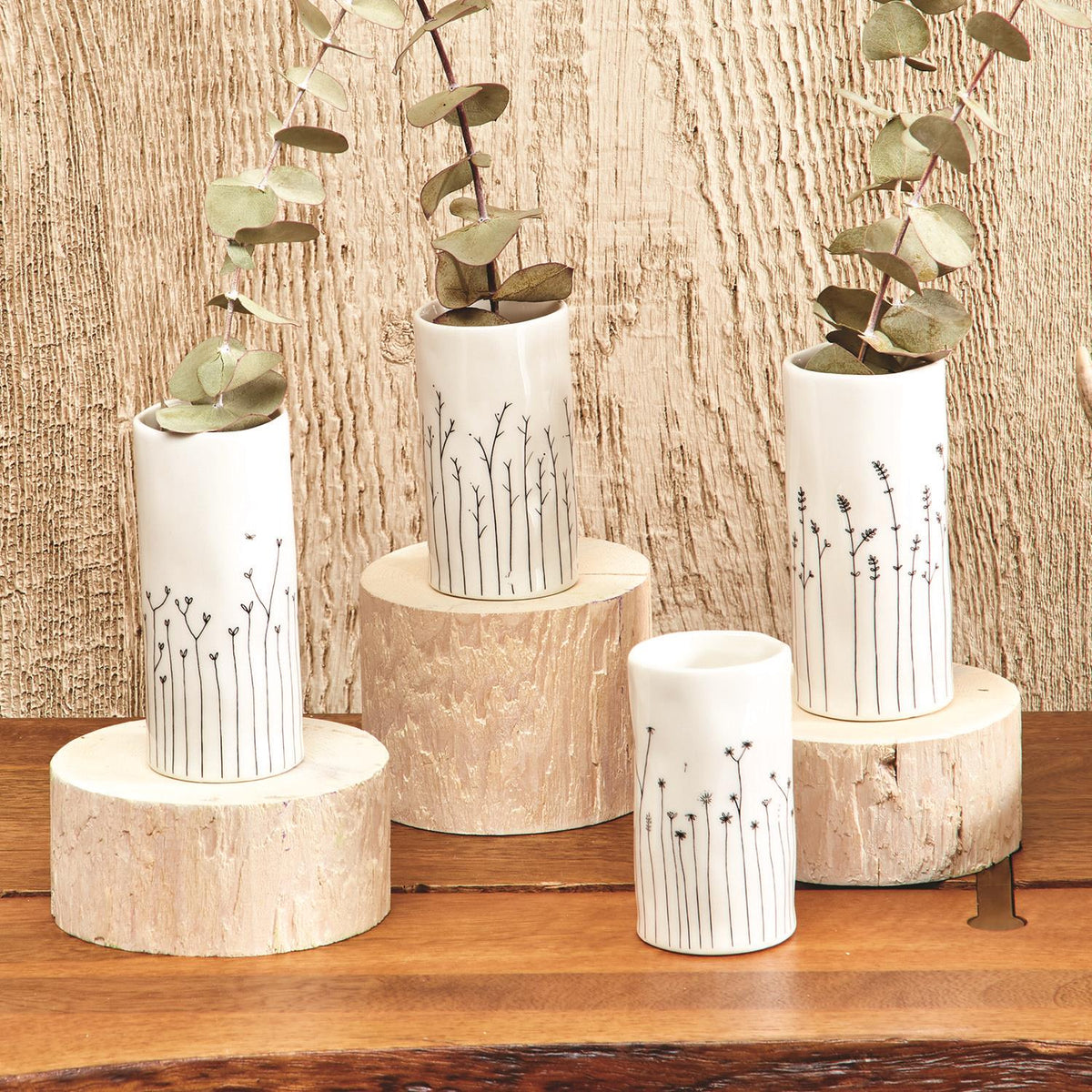 White porcelain mini vases with hand-drawn stem designs holding eucalyptus sprigs on wood slice risers