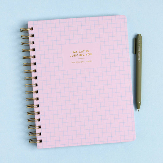 Lavender grid spiral notebook with gold foil title, My Cat Is Judging You, beside a green pen