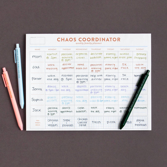 Chaos Coordinator weekly family planner pad filled in, with colorful pens on a dark surface